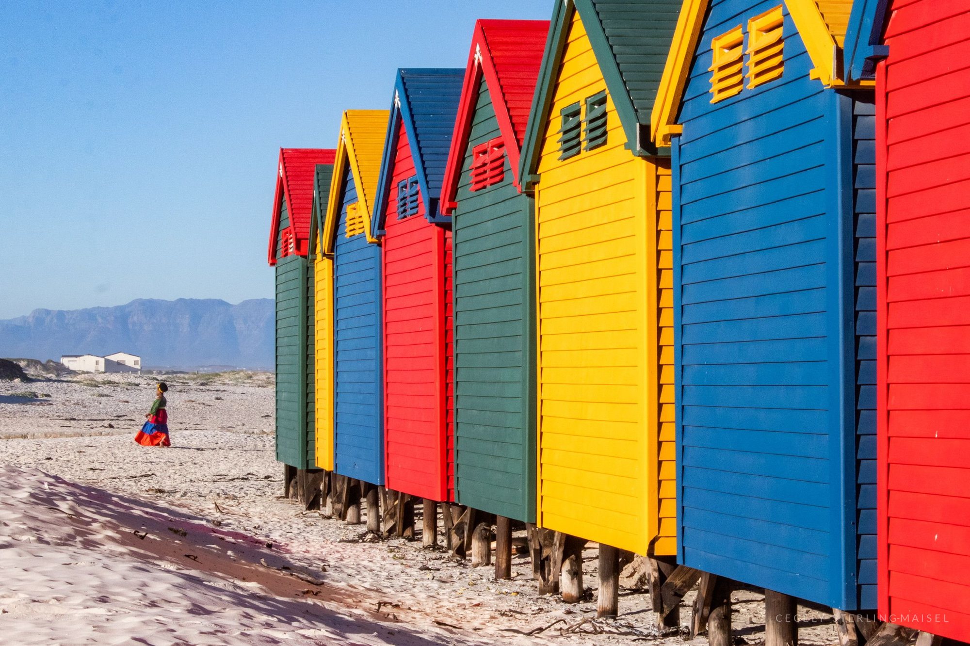 A row of red, yellow and blue beach huts on a pale beach, one figure in red walking past them.