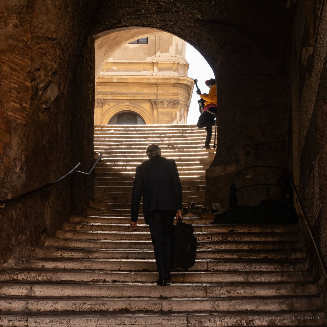 A man with a suitcase climbing a wide stone staircase toward a sunlit baroque facade, seen through a stone arch.