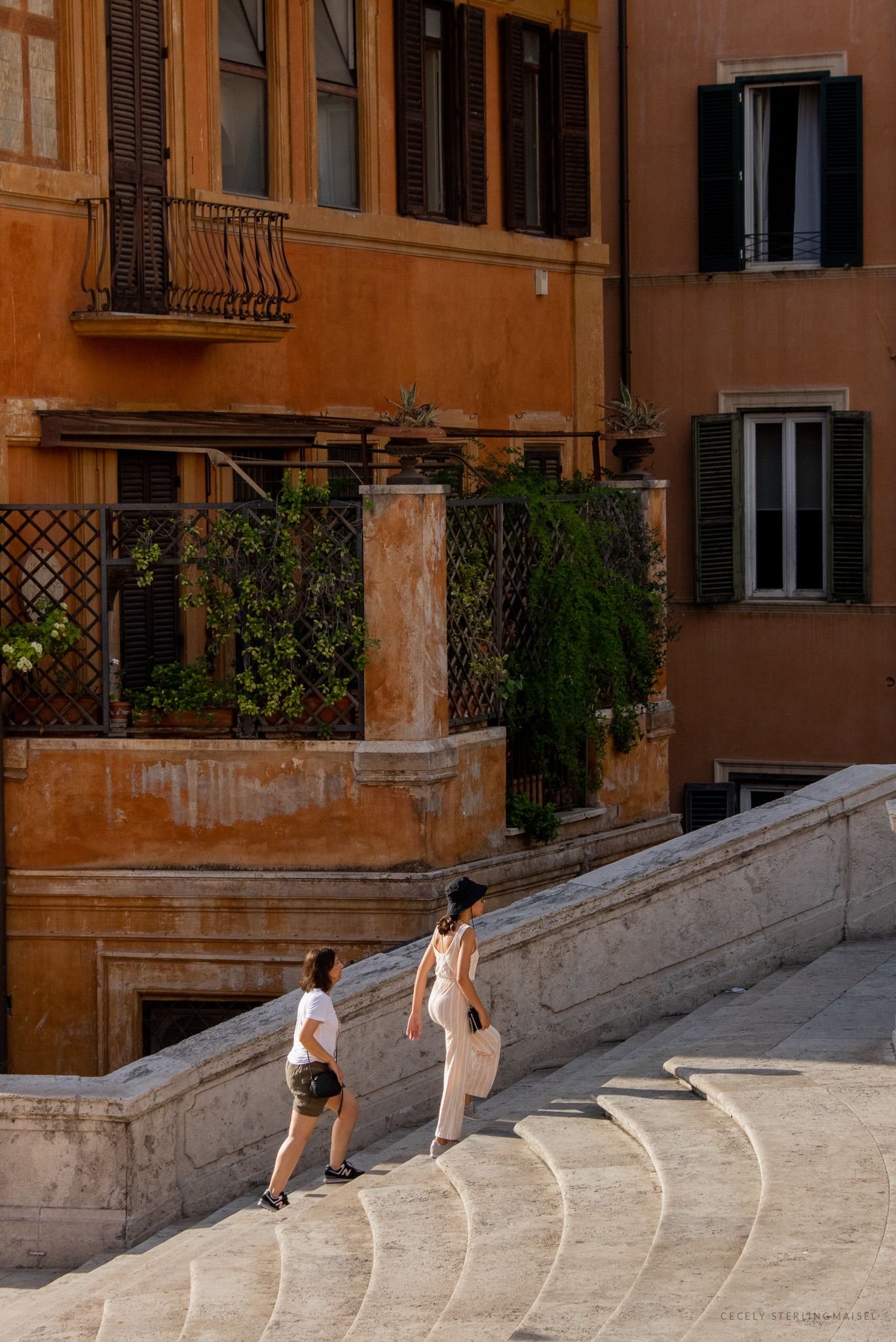 Terracotta buildings with green shutters above a curving flight of steps where two figures pass.