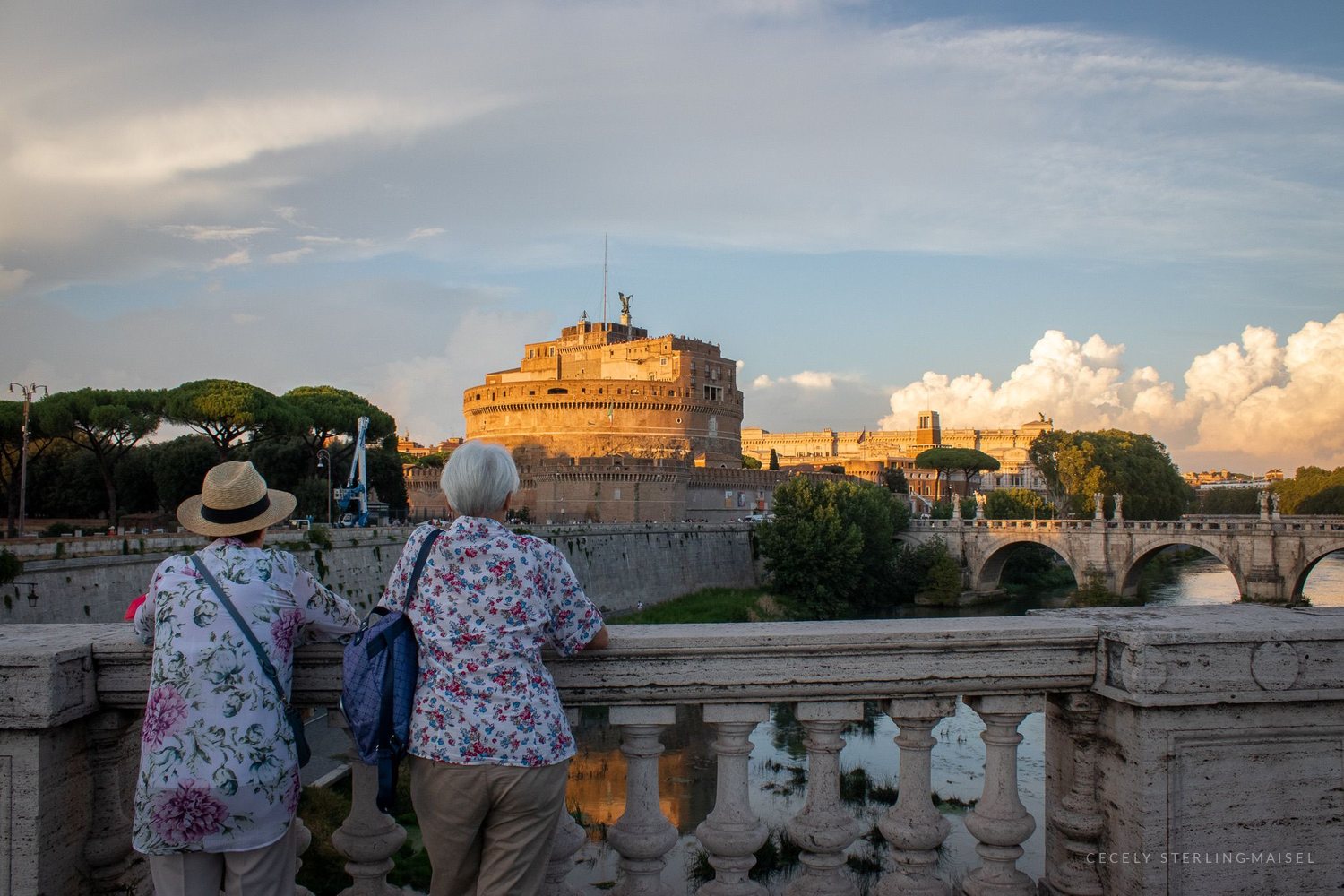 Two women in sun hats leaning on a stone balustrade looking toward Castel Sant'Angelo at golden hour.