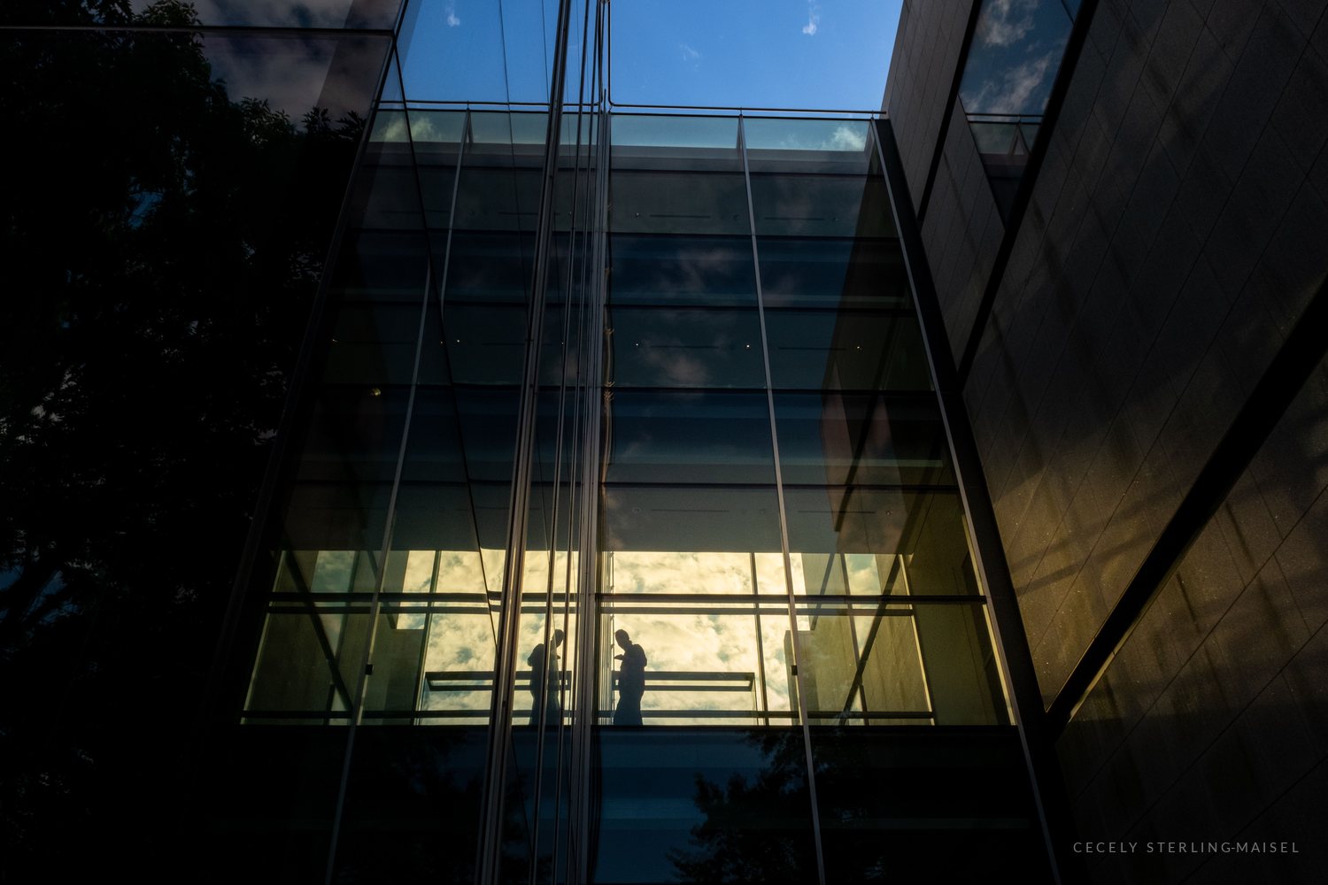 Looking up at a dark glass and stone building where two silhouetted figures stand at a railing against a bright, cloud-filled window.