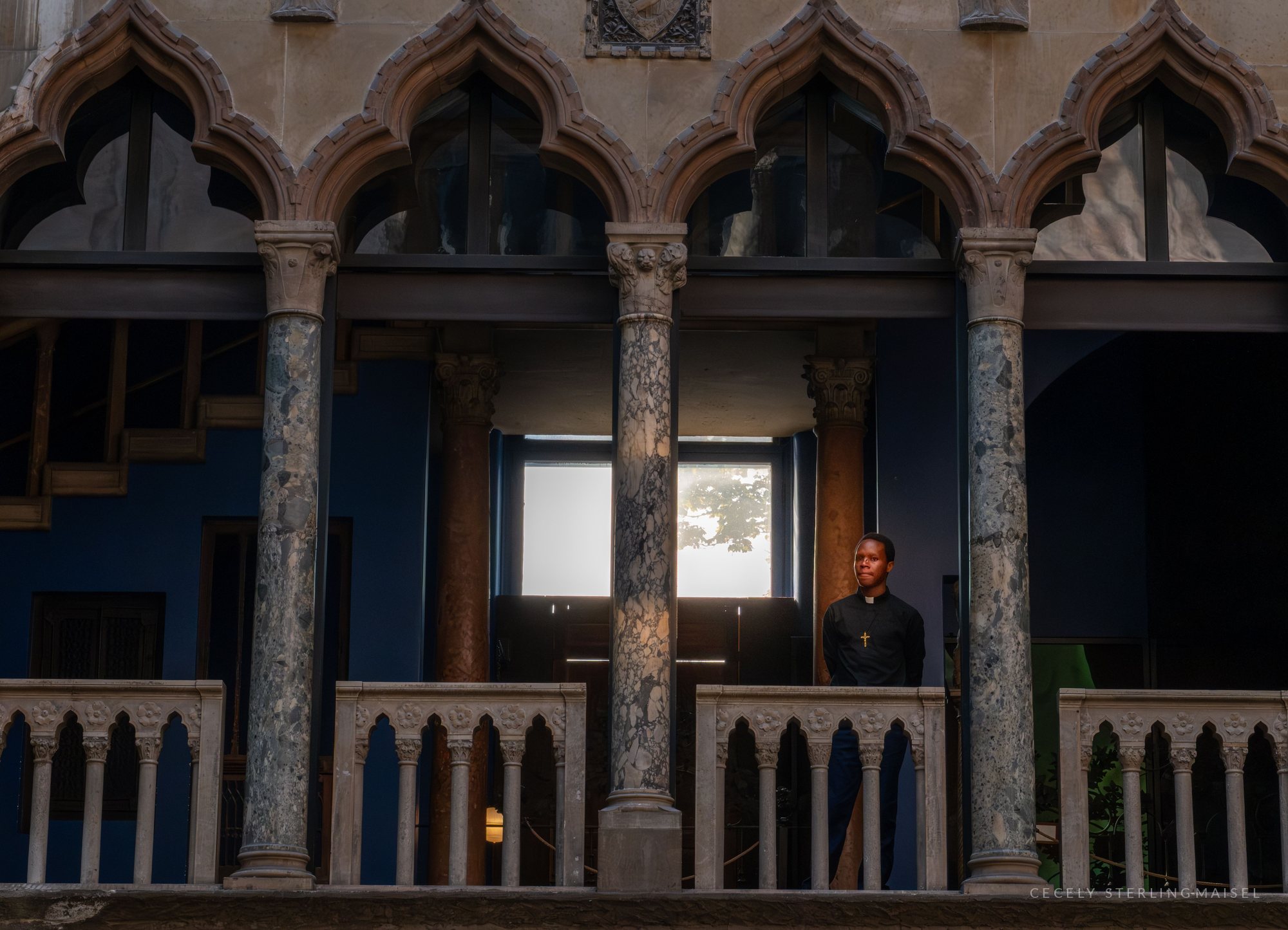 A person in dark clerical clothing with a cross stands on a balcony behind an ornate Gothic arcade of carved columns and pointed arches.