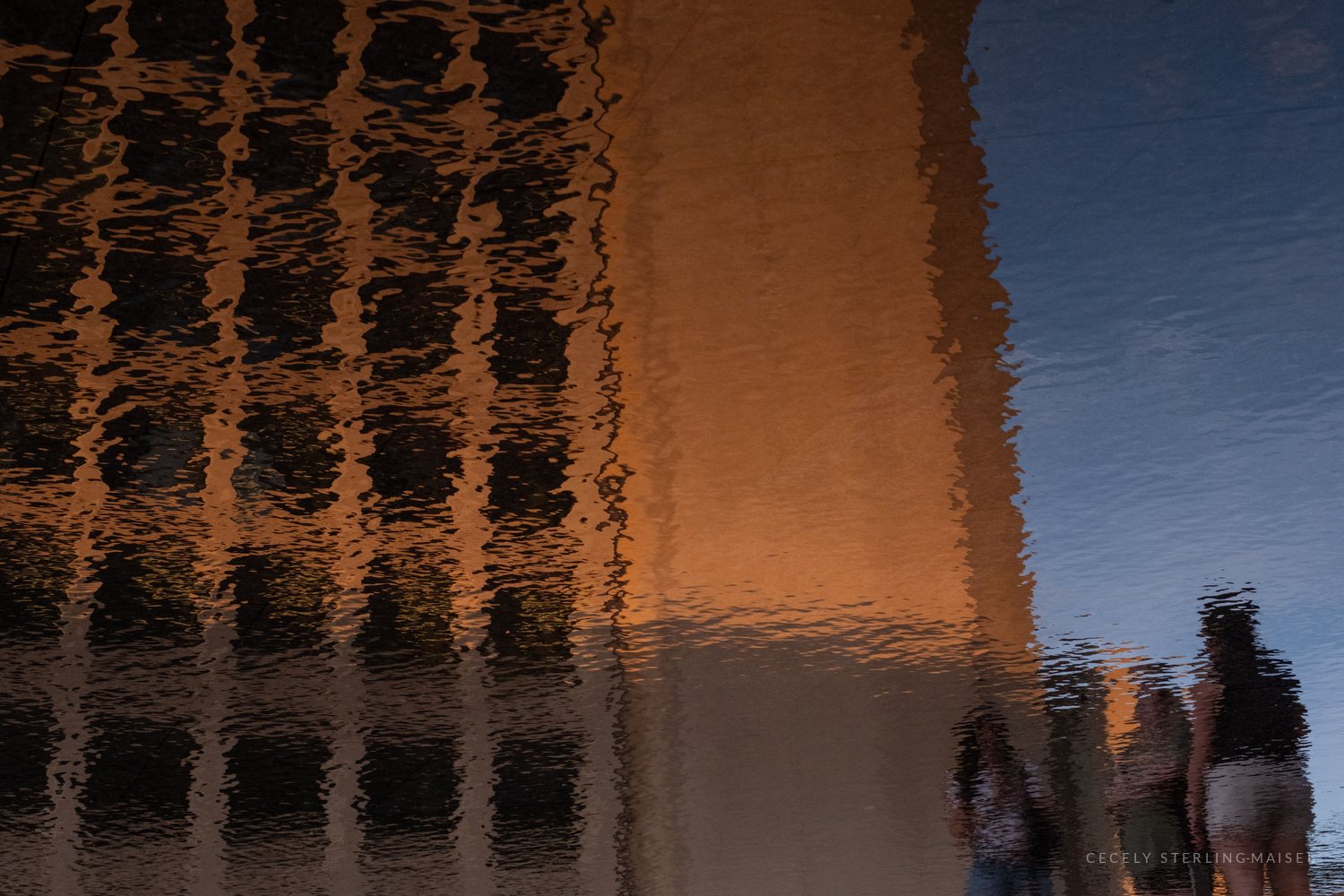 Rippling water reflecting a brick building glowing orange at sunset, with the wavering silhouettes of three people standing at the edge.