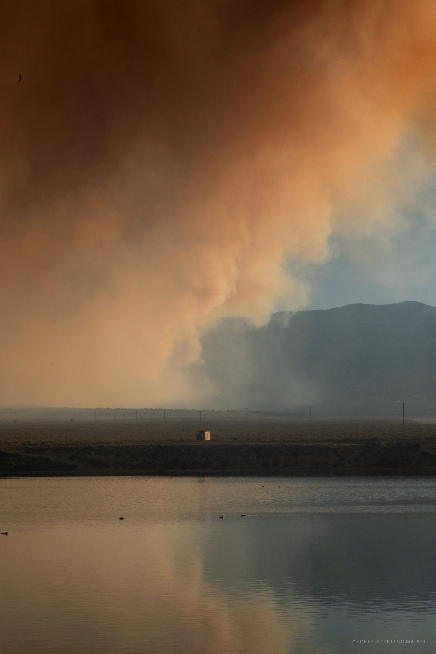 A column of wildfire smoke lit orange by the sun rises above a reservoir, with one small lit building on the far shore.