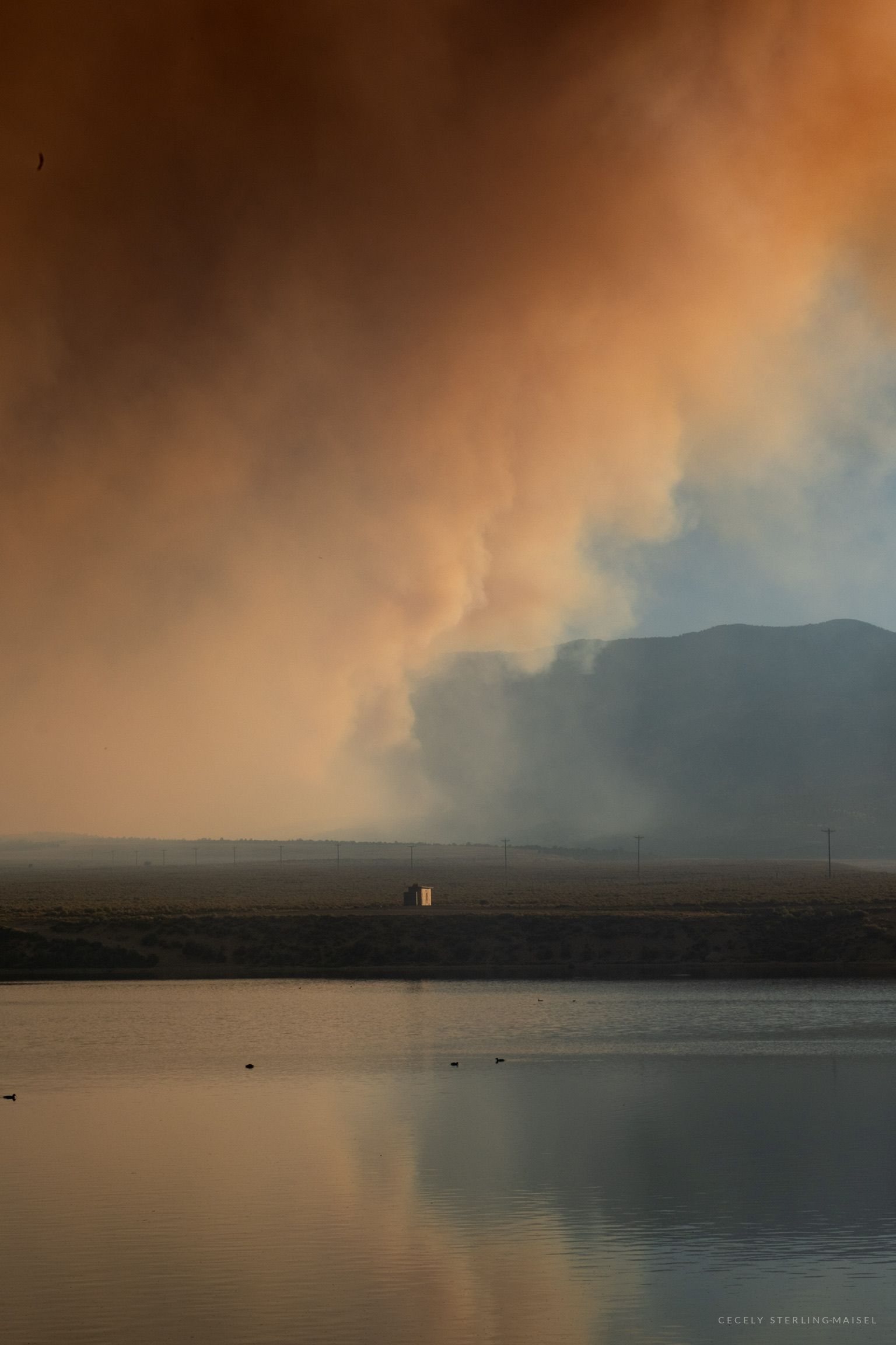 A column of wildfire smoke lit orange by the sun rises above a reservoir, with one small lit building on the far shore.