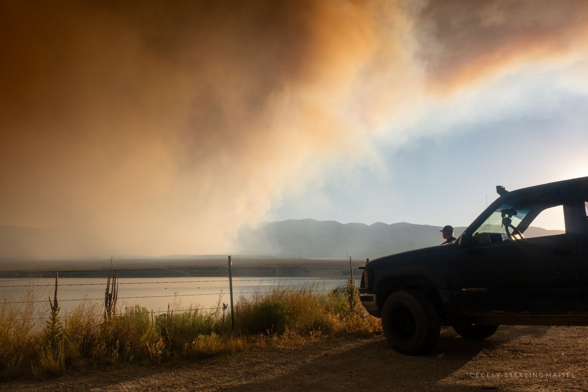 A person stands beside a pickup truck at the roadside watching wildfire smoke pour across the sky above a reservoir.