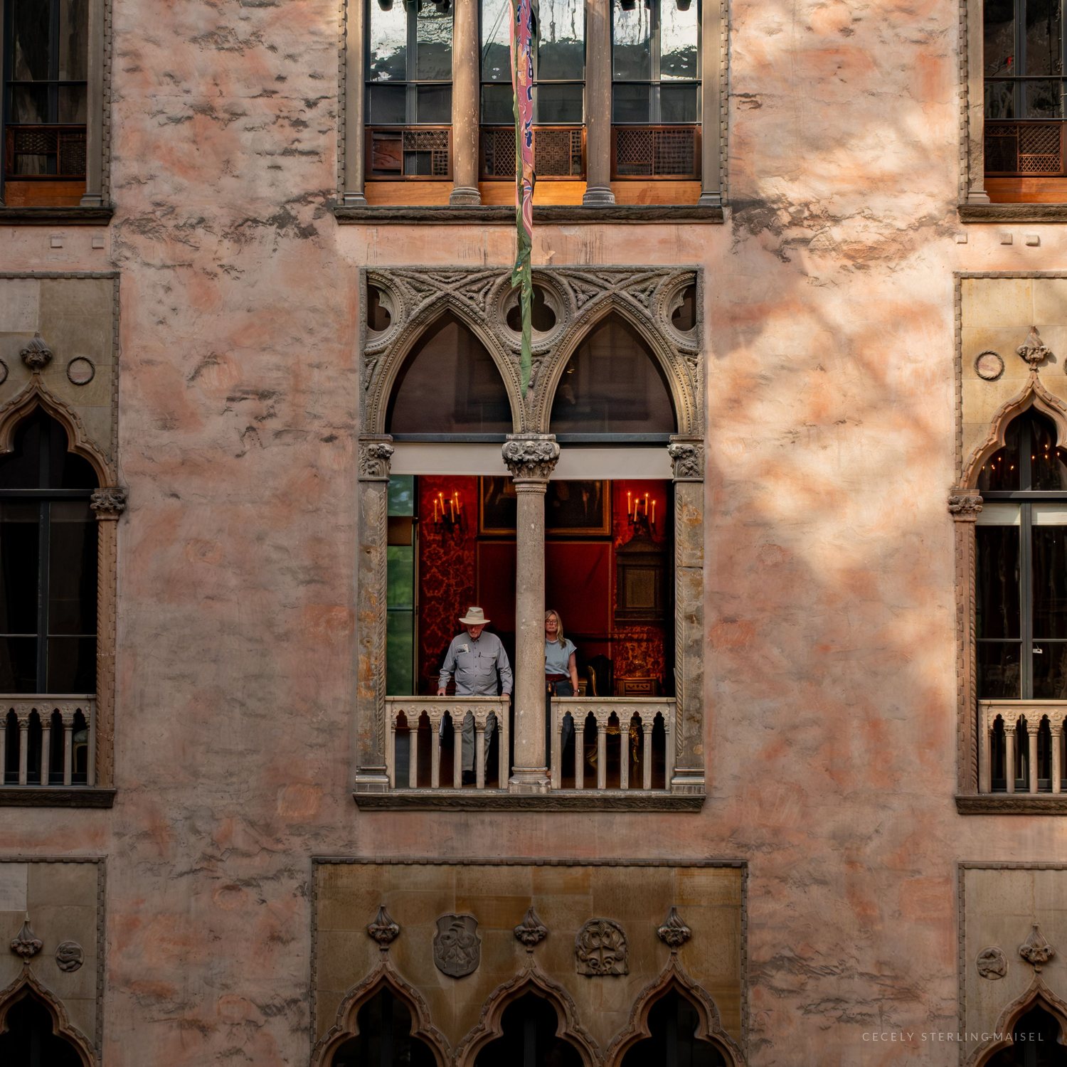 Two visitors lean on the railing of a Venetian-style arched window in a pink stone courtyard wall, a candlelit red room glowing behind them.