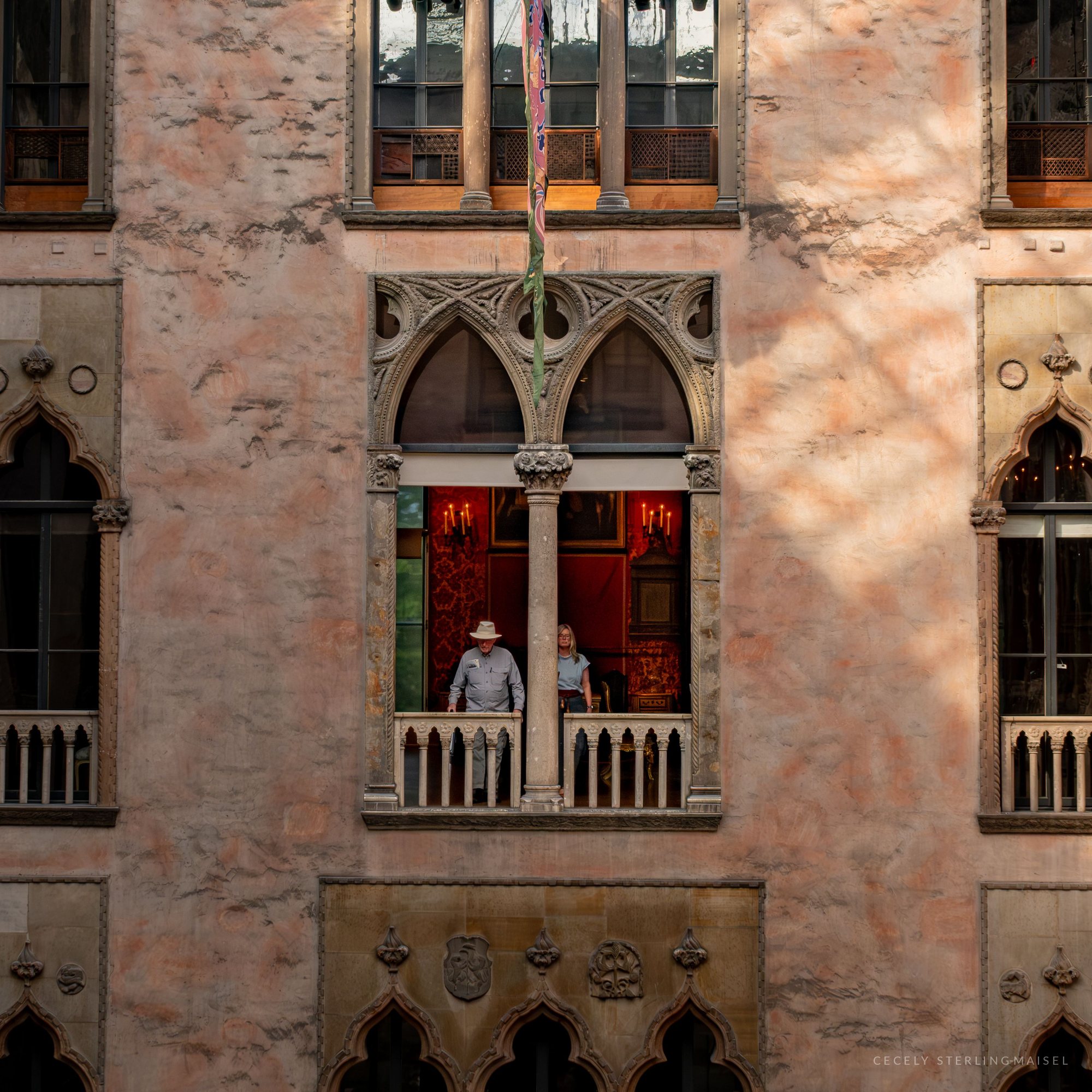 Two visitors lean on the railing of a Venetian-style arched window in a pink stone courtyard wall, a candlelit red room glowing behind them.