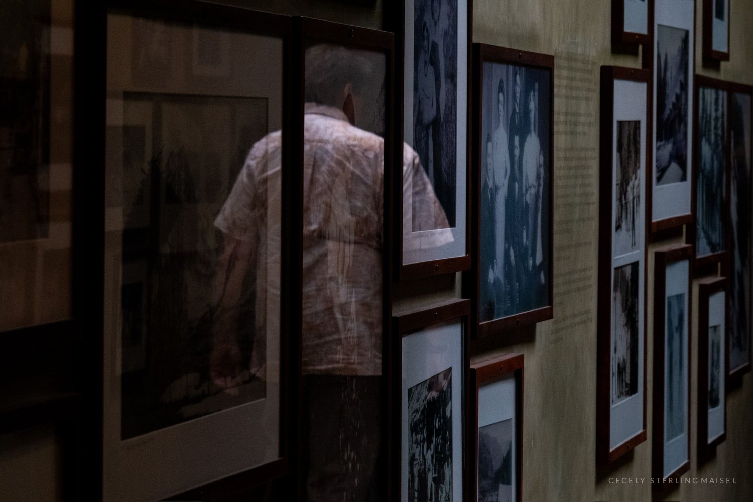 A man in a pale short-sleeved shirt, seen as a reflection in the glass of a large framed picture among a crowded gallery hang.
