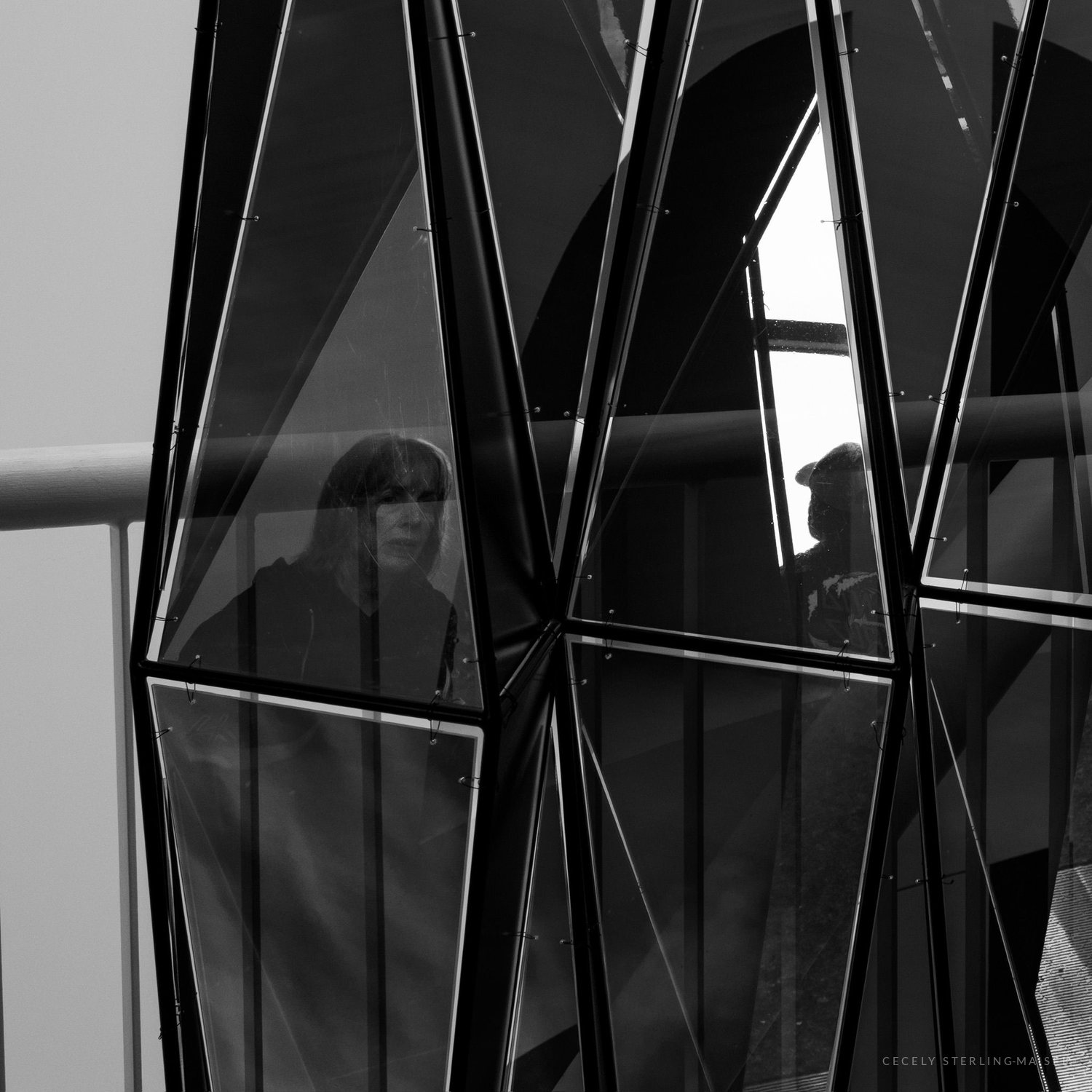 Black and white: a woman stands behind the faceted glass panels of a building, her face caught between the diagonal frames.