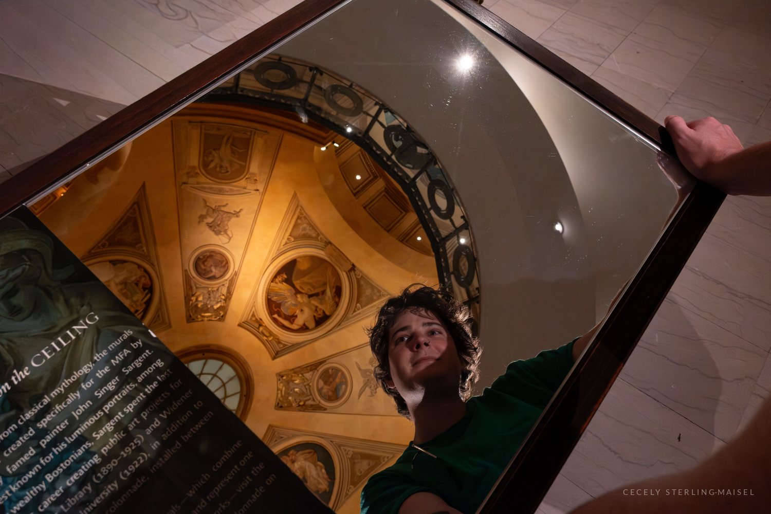 A young man holds up a large handheld mirror that reflects both his face and an ornate painted museum ceiling above him.